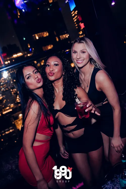 Three women posing together at a nighttime party organized by 1990 Group in New York, one of them holding a drink.