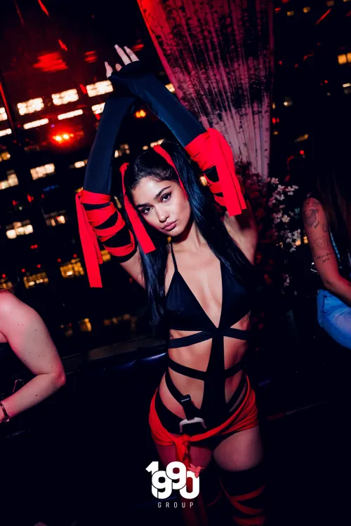 Woman posing in a red and black outfit, with her hands raised at a themed party organized by 1990 Group in New York.