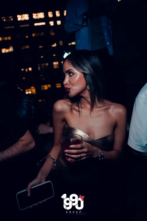 Elegant woman in a gold dress enjoying a party organized by 1990 Group in New York, with the city in the background.