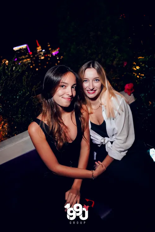 Two friends posing at a nighttime party organized by 1990 Group in New York, with city lights in the background.