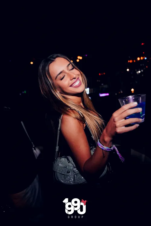 Smiling woman with a drink in her hand at a party organized by 1990 Group in New York, a fun nighttime atmosphere.